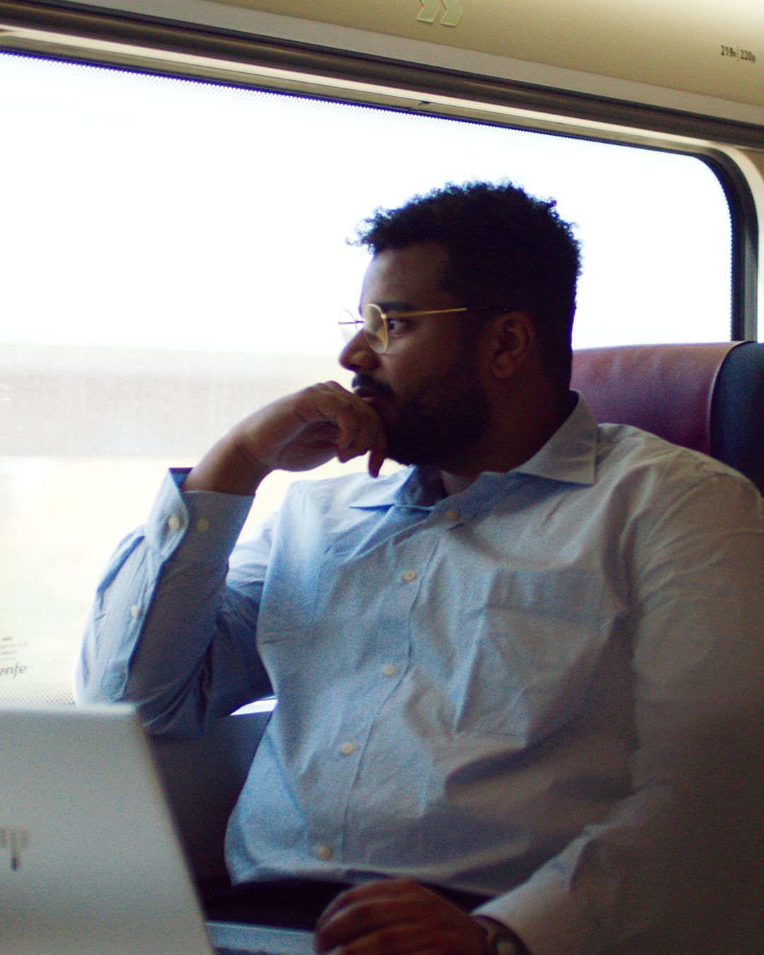A man in glasses gazes out the train window, his laptop in front of him